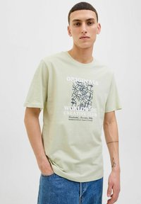 Lys grøn bomulds-T-shirt med korte ærmer, der har et grafisk design med sorte linjer og tekst. Løs pasform, parret med blå denimjeans.