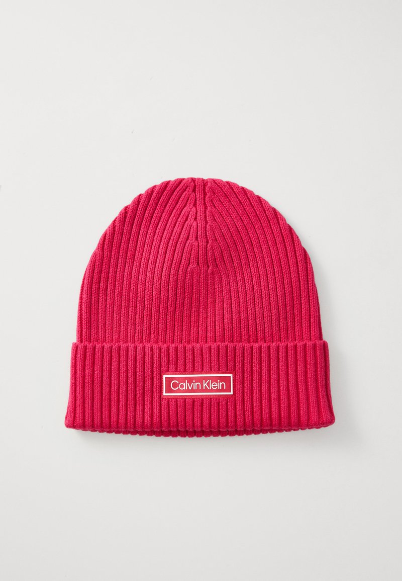 Růžová pletená čepice s ribováním a ohnutým lemem, na které je obdélníková bílá značka se červeným okrajem s nápisem "Calvin Klein."