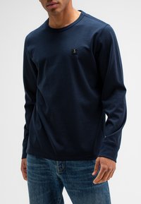 Marineblaue Langarmshirt aus weichem Stoff, mit Rundhalsausschnitt und kleinem Logo-Patch auf der Brust. Kombiniert mit Jeans.