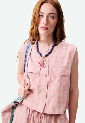 Jonge vrouw met lang rood haar, gekleed in een mouwloos roze geruit topje en een ketting met paarse kralen en een roze kreeftenhanger.