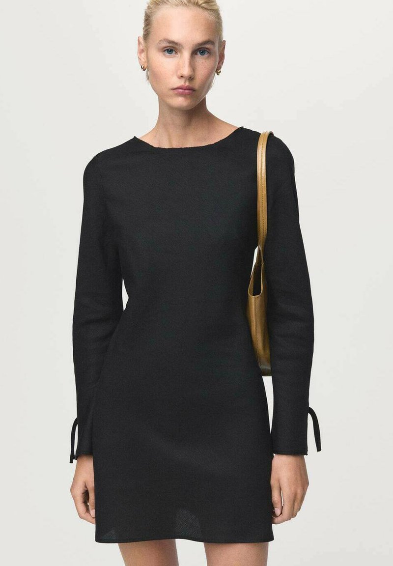 Mujer con cabello rubio que lleva un simple vestido negro de manga larga y lleva un pequeño bolso de hombro color beige sobre un fondo claro.