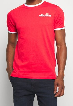 Mand iført en knaldrød kortærmet t-shirt med hvid kant og lille hvid ellesse-logo på brystet, kombineret med mørke jeans.