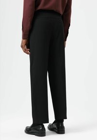 Pantalon noir à jambes larges en tissu lisse, avec devant plat, deux poches arrière et une coupe ajustée. Porté avec des chaussures noires.