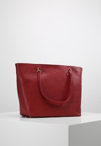 Czerwona skórzana torba tote o teksturowanym wykończeniu, z dwoma uchwytami i metalowymi akcesoriami, wyeksponowana na jasnym podwyższeniu.