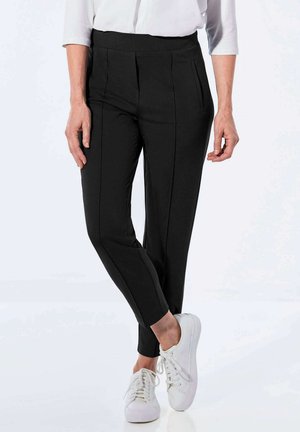 Pantalons noirs ajustés avec des plis, des poches latérales et un design épuré. Associés à des baskets blanches pour un look décontracté.