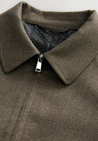 Manteau en tissu marron texturé avec un col pointu et un tirette de fermeture éclair argentée visible sur une doublure noire à motif.