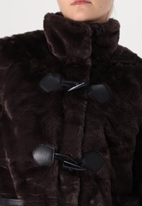 Brązowy płaszcz z futerka ekologicznego z wysokim kołnierzem, z czarnymi zapięciami na klamry i gładkimi skórzanymi wstawkami. Miękka faktura i krótki fason.