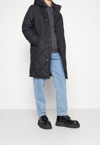 Czarna pikowana kurtka puchowa z kapturem, z wzorem w romby, noszona na szarej bluzie z kapturem, w połączeniu z jasnoniebieskimi dżinsami i czarnymi masywnymi butami.