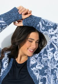 Modrý a bílý cardigan s vzorem, s žebrovanými manžetami a otevřeným předním dílem, vyrobený z měkké texturové látky. Model předvádí uvolněnou pozici.