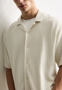Light beige short-sleeve shirt with a textured pattern, button closure, and a collared neckline, worn over a plain white undershirt.