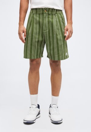 Mand iført grønne stribede shorts, hvide sokker, hvide sneakers og en hvid skjorte, stående mod en lys baggrund.