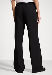 Osoba v černých kalhotách s širokými nohavicemi a černých botách, stojící zády k fotoaparátu na jednoduchém bílém pozadí.