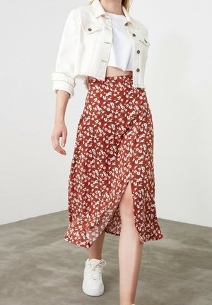 Vrouw draagt een witte korte jas, wit crop top, rode midi-rok met bloemenprint en een voorste split, en witte sneakers, wandelend op een grijze vloer.