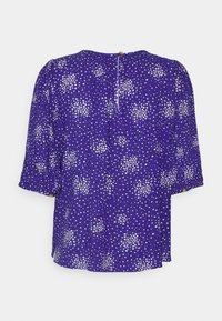 Blusa roxa com padrão de bolinhas brancas, mangas curtas bufantes, fecho de botão nas costas e um corte descontraído com a barra ligeiramente curvada.