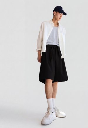 Jeune homme debout portant un short large noir, un débardeur blanc, une chemise blanche ouverte, des baskets blanches, des chaussettes et une casquette de baseball bleu marine sur fond uni.