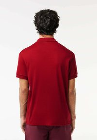 Rød polo-shirt lavet af glat stof, med en klassisk krave, korte ærmer og en lige nederkant, vist fra bagsiden.