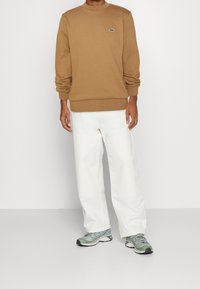 Sudadera marrón con puños y dobladillo acanalados, pantalones anchos blancos y zapatillas deportivas verdes y grises. Logotipo de Lacoste en el pecho.