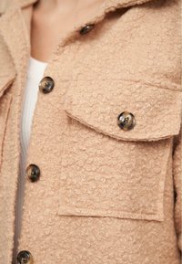 Beige texturovaná bunda s chlupatým povrchem, velkou náprsní kapsou a kulatými knoflíky. Obsahuje límec a mírně oversize střih.