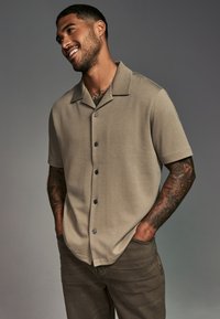 Korte mouwen, knoopshirt van lichtbeige stof met kraag en donkere knopen, gecombineerd met een donkerder beige broek. Gladde textuur.