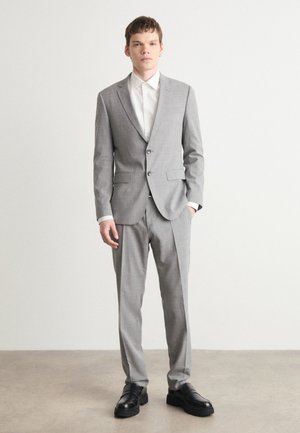Grau karierter Anzug mit zweiknöpfigem Blazer und flachen Hosen. Weißes Hemd darunter, kombiniert mit schwarzen Leder-Schuhen. Glattes Material und taillierte Passform.