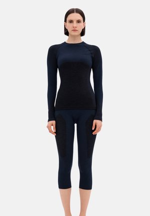 Frau steht frontseitig, trägt ein figurbetontes schwarzes Langarmoberteil und passende kurze Leggings vor einem schlichten hellen Hintergrund.