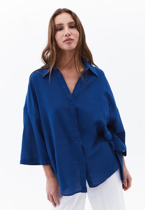 Blue oversized button-up shirt with a textured grid pattern, collar, and wide sleeves. Paired with light-colored trousers.