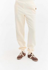 Kremowe spodnie dresowe z elastycznym pasem. Posiadają boczne kieszenie i gumowy akcent. W połączeniu z brązowymi trampkami i białymi sznurowadłami.