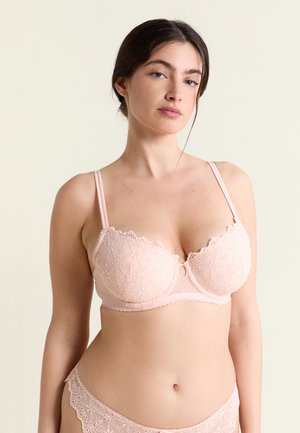 Femme portant un soutien-gorge en dentelle florale rose clair et une culotte assortie, debout devant un fond beige uni, faisant face à l'avant.