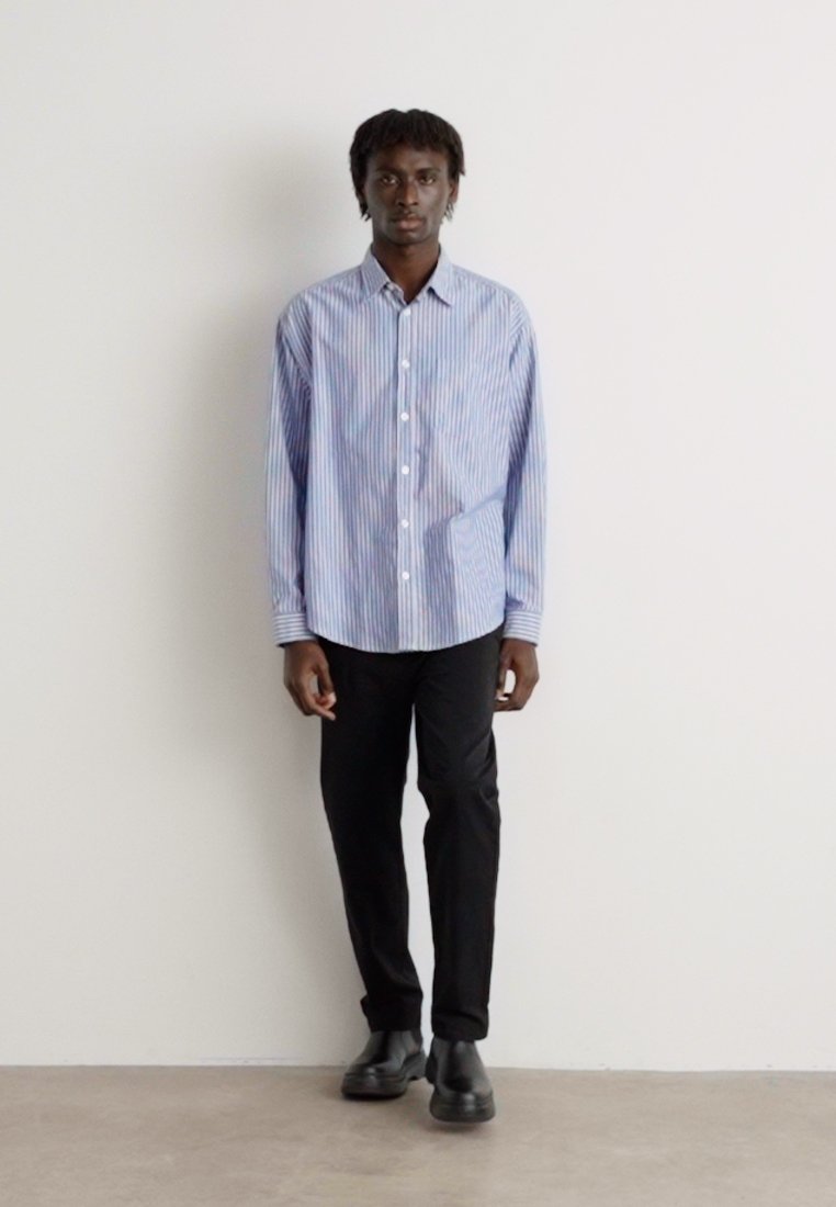 Chemise à manches longues à rayures bleues et blanches, associée à un pantalon noir et des chaussures noires, sur fond neutre.