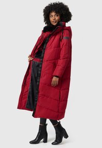 Long red puffer coat with a faux fur collar, featuring a zipper and snap buttons, black lining, and side pockets. Worn over black pants and boots.