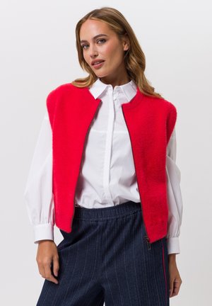Rood, pluizig mouwloos vest met een hoge halslijn en ritsluiting, gedragen over een wit shirt; gecombineerd met donkerblauwe broek met fijne krijtstrepen.