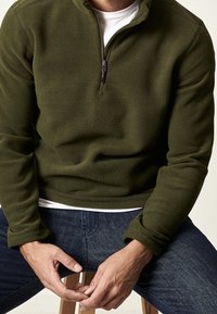 Olivengrüner Reißverschluss-Fleece-Pullover mit Kragen und langen Ärmeln, kombiniert mit dunkelb blauen Jeans. Weicher Stoff und lockere Passform.