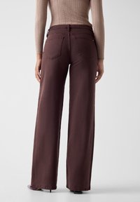 Jeans a gamba larga e vita alta in denim marrone scuro, con due tasche posteriori, cuciture minimali e una finitura liscia e strutturata.