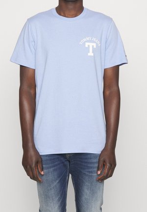 Mężczyzna w jasno-niebieskim t-shircie Tommy Jeans z białym logo na lewej stronie klatki piersiowej i wyblakłych niebieskich dżinsach, stojący na jednolitym tle.