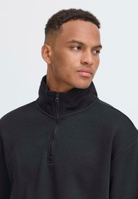 Sweatshirt noir zippé avec col haut. Présente une texture lisse et une demi-fermeture éclair à l'avant. Design simple, sans motifs ni accents.