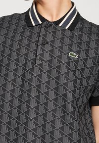 Personne portant un polo noir à motifs avec un col noir et des rayures blanches, avec un petit logo crocodile vert sur la poitrine.