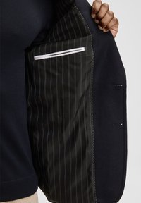 Wnętrze czarnej kurtki z podszytą kieszenią w niebieskie paski. Tkanina wygląda na gładką, o jednolitej fakturze i widocznych detalach przeszyć.