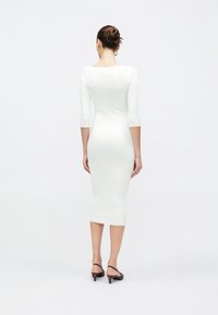 Γυναίκα με τα μαλλιά της σε κότσο, που φοράει εφαρμοστό, λευκό μίντι φόρεμα με ριγέ υφή και μανίκια τριών τετάρτων, καθώς και καφέ χαμηλά πέδιλα με λουράκι πίσω, κοιτάζοντας προς τα πίσω.