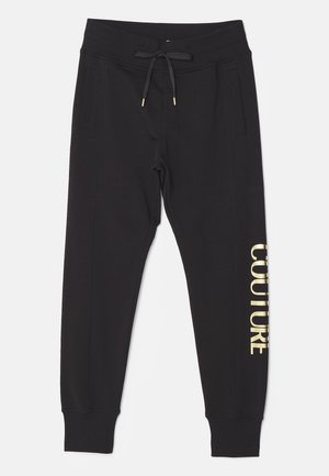 Homme portant un sweat-shirt noir et un pantalon avec le texte doré « Versace Jeans Couture », assortis à des chaussures de sport noires et blanches.