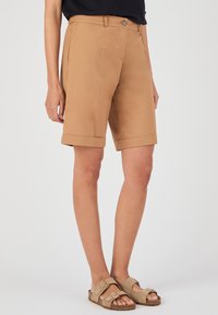 Pantalones cortos beige con frente plano, las pasadores para cinturón y bajos enrollados. Hechos de tejido suave. Se usan con sandalias tipo slipper de color marrón claro.