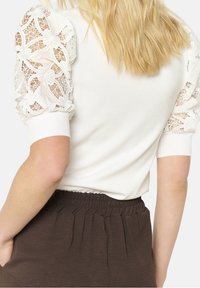 Vit nära sittande topp med genomskinliga spetspuffärmar som visar upp intrikata mönster. Baksidan är slät och kombineras med byxor med brun elastisk midja.