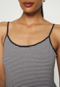 Mujer vistiendo una camiseta sin mangas de rayas horizontales en blanco y negro con tirantes delgados y pequeños pendientes de aro dorados, sobre un fondo blanco.