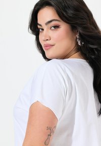 T-shirt bianco a maniche corte con una vestibilità rilassata. Presenta un tessuto morbido, un collo a giro e un tatuaggio visibile sul braccio di chi lo indossa.