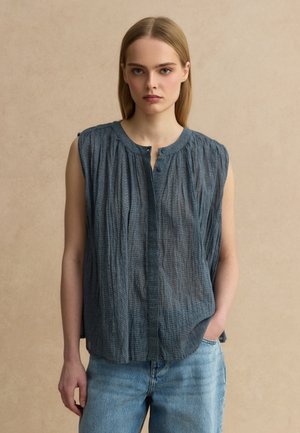 Junge Frau mit glattem, blondem Haar trägt eine ärmellose, dunkelgraue, plissierte Bluse und hellblaue Jeans und steht vor einem beigen Hintergrund.