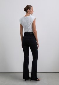 Kvinna med hår i en bulle, iförd en vit texturerad ärmlös topp, höga svarta jeans och svarta klacksandaler, stående mot en grå bakgrund.