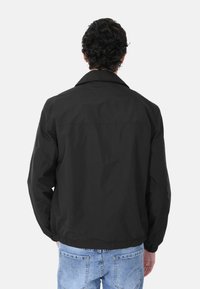 Giacca leggera nera con colletto, tasche laterali e polsini elastici. Tessuto liscio e vestibilità comoda, abbinata a jeans in denim blu.