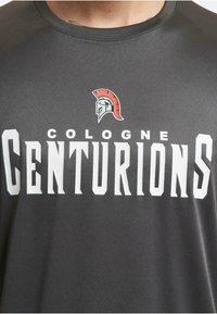 Pilka trumpa rankovė marškinėliai su ryškiu baltu užrašu, kuriame parašyta "KOLONIJOS CENTURIONAI", ir emblema su spartiečių šalmą viršuje.