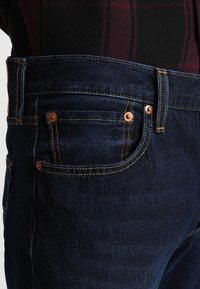 Vaqueros de mezclilla azul oscuro con remaches de cobre, costuras naranjas y un diseño clásico de cinco bolsillos. Textura suave con un ligero estiramiento.