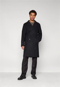 Μαύρο διπλόφερμο παλτό με φαρδιούς γιακάδες, κατασκευασμένο από υφή ύφασμα. Διαθέτει μπροστινές τσέπες και φοριέται πάνω από γκρι πουλόβερ και παντελόνια.