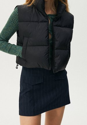 Frau trägt eine schwarze gesteppte Weste über einem grünen, gerippten Pullover und einem marineblauen Nadelstreifenrock mit vorderen Klappentaschen.